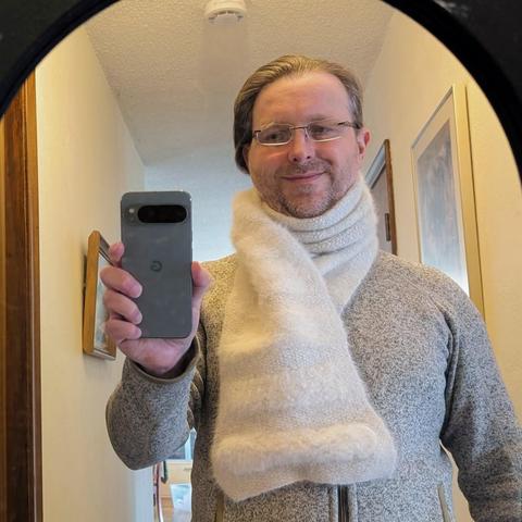 A guy takes a selfie photo in a mirror in a hallway, showing off his hand-knit husky yarn scarf. It's wrapped around his neck in a thick coil up to his chin, and one end lays flat over his chest, showing off sections of extra fluffy yarn.