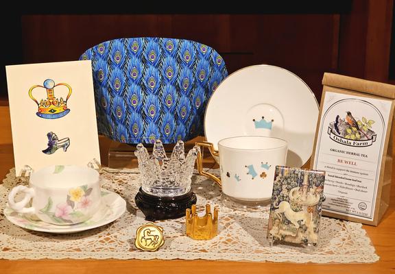 Tea tableau with a peacock feather pattern tea cozy, a crystal crown, a gold crown, a gold medallion showing a unicorn, a small picture of a Bayeux Tapestry detail of a unicorn, a card with a crown and fancy slippers, vintage china, and  herbal wellness tea from Oregon.
