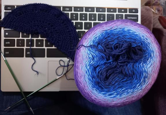 Photo shows my newest knitting WIP, a teeny tiny semi circle of dark blue yarn, with the ball km knitting from beside it, a gradient yarn going from blue to purple.