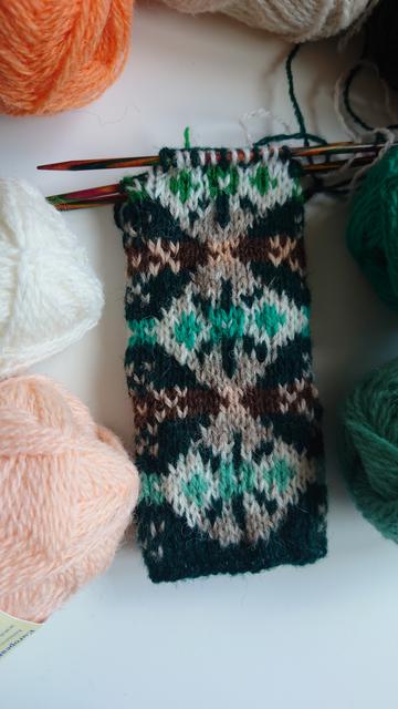 Show a knitted tube in dark bottle green with pale diamond shapes in traditional Fair Isle patterns. The diamonds have a green blue centre pattern section starting in mint, then the next has jade, and the final one has celtic green. The swatch is still on the needles, and lying on a white table surrounded by the other balls of wool used. It's under a natural light.