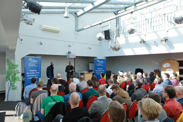 Two male persons standing on a stage between two FSFE roll-ups and talking to an audience