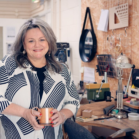 A woman holding a copper mug is leaning against a jeweller's workbench. She is holding a copper mug, and wearing a dress with a stripy patten. On the bench is an angle-poise lamp and various tools associated with making silver jewellery. Hanging on the wall behind are coils of wire and a template for drawing angles and circles. Behind the bench can be seen some power tools.