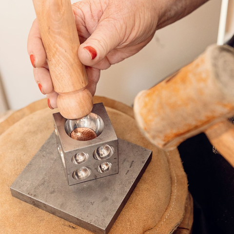 Circle of copper being formed into a dish shape in a metal die. The die resembles a large steel die with exaggerated indentations. There is a hand holding a wooden form for pressing the copper into the metal die, and the head of a rawhide hammer is blurred in the foreground.