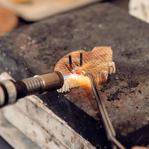 Blowtorch being used to solder a jump ring onto a copper pebble. The pebble is held in place by two prongs placed in a heatproof grid, and the jump ring is being positioned using tweezers.
