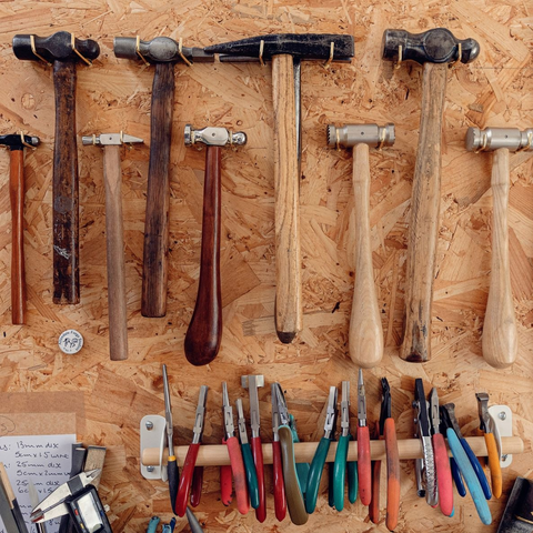 Selection of nine hammers of varied shapes and sizes hanging on a piece of board. Below the hammers are about a dozen assorted pliers of different styles, hanging on a piece of dowel. The end of a digital caliper can also be seen.