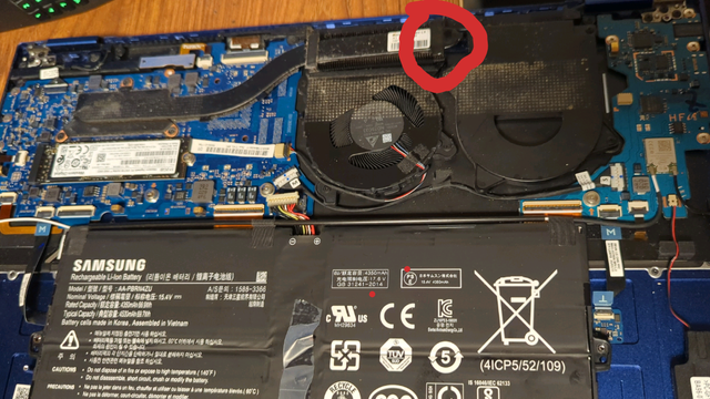 A picture of the inside of a laptop. It's very blue colored & has a disappointingly small battery. One of the two fans is actually fake, despite there being a plug for a second fan on the sub motherboard. The blob of black tar looking glue on the heat pipe is circled in red. The heat pipe is very long with nothing securing it except its connection to the CPU, all the way on the opposite end & opposite side from the glue! This causes a lever action when you pull on the bottom casing panel, bending the heat pipe & causing a pinch in it near the CPU.