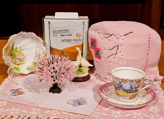 Tea tableau in pink with a paper cutout cherry tree in full bloom at center, a cherry blossom tea cozy, ornately floral vintage china, pastel painted butterflies, and a pair of bisque porcelain snow geese, on a pink and white tea towel. The tea is Cherry Marzipan Oolong loose leaf from Adagio.